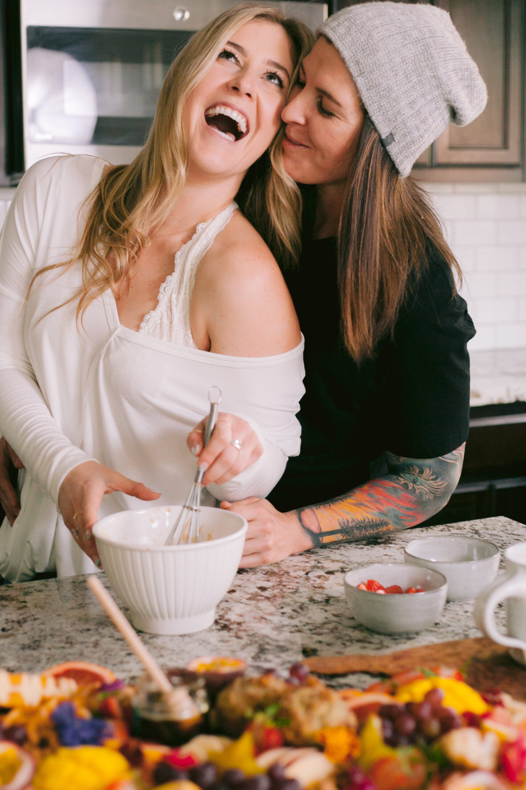 Woman hugs woman from behind in kitchen. 
