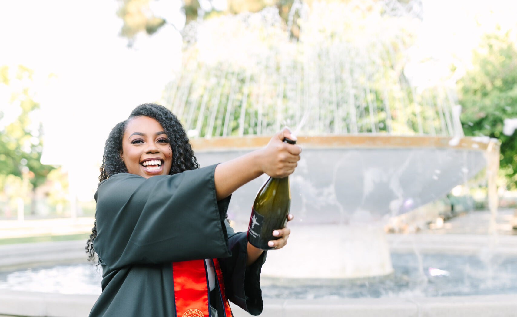 Girl in black graduation gown popping champagne bottle infront of fountain at Fresno State taken by GunnShot Photography